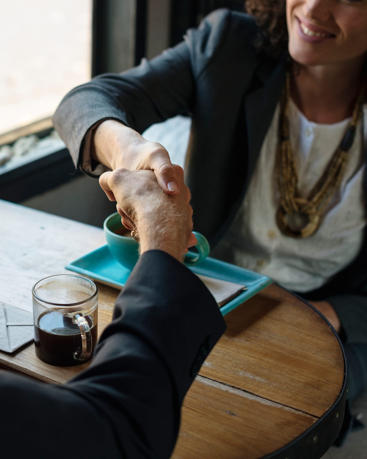 Two professionals in dark jackets shaking hands across a wooden cafe table with coffee cups