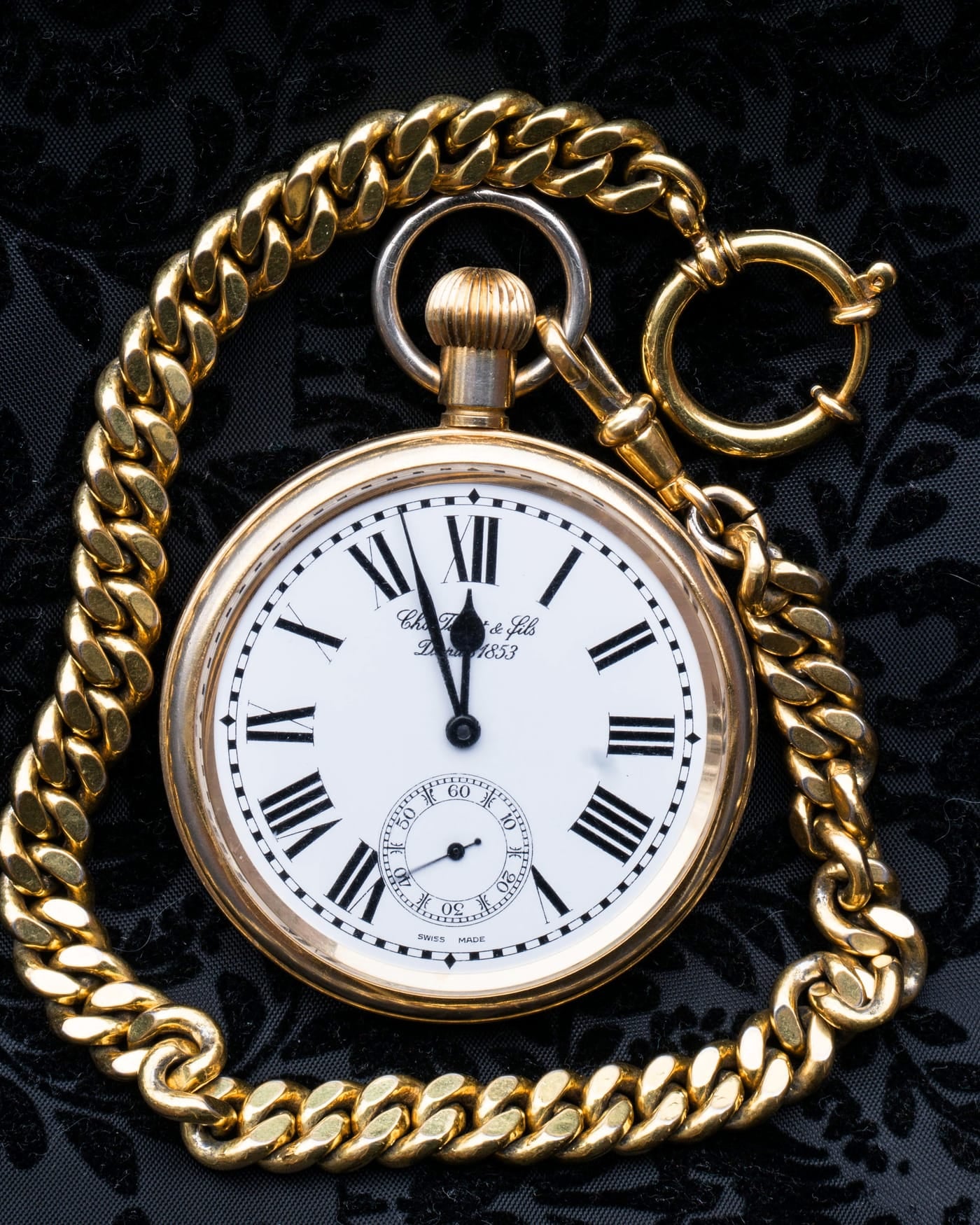 Gold pocket watch with a chain resting on dark patterned fabric