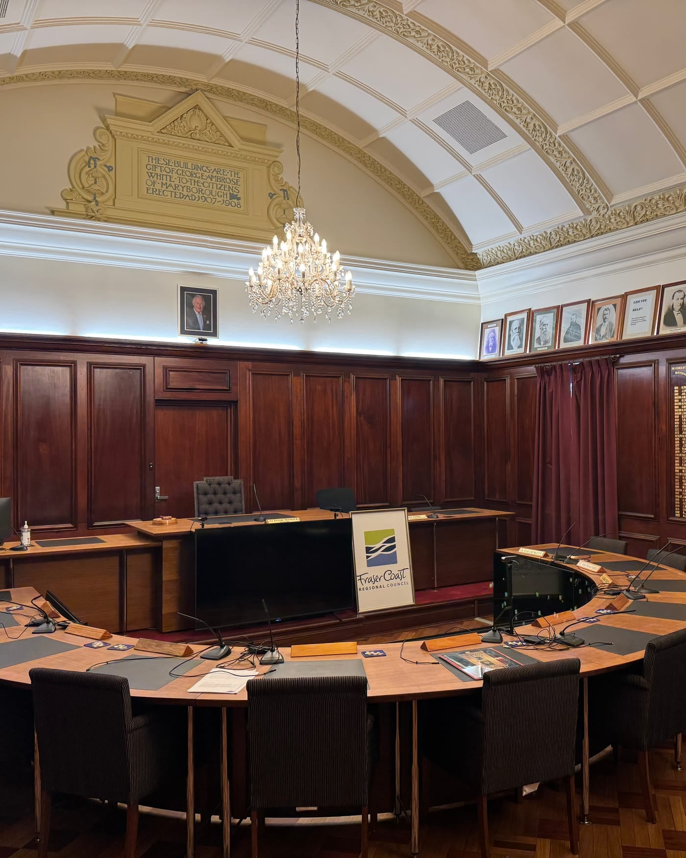 Empty council chambers with a curved wooden desk, chairs and a chandelier