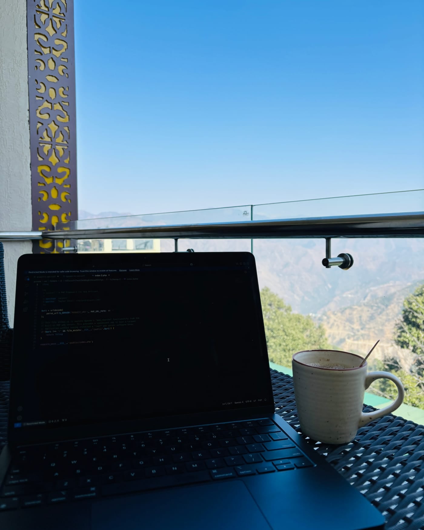 Laptop with a code editor open on a balcony table beside a mug, with blue sky and mountains behind
