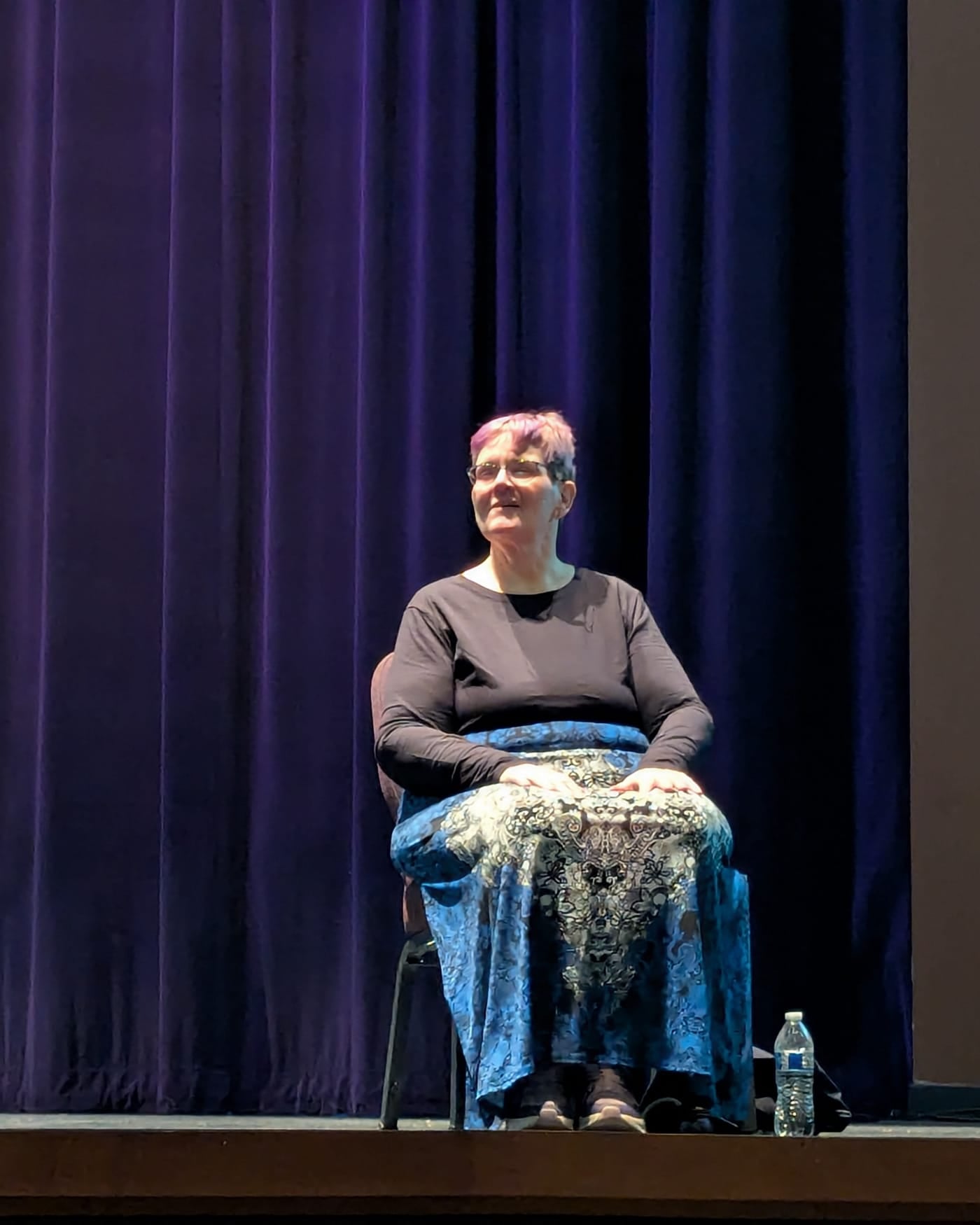 Sign language interpreter seated on a stage in front of a dark blue curtain