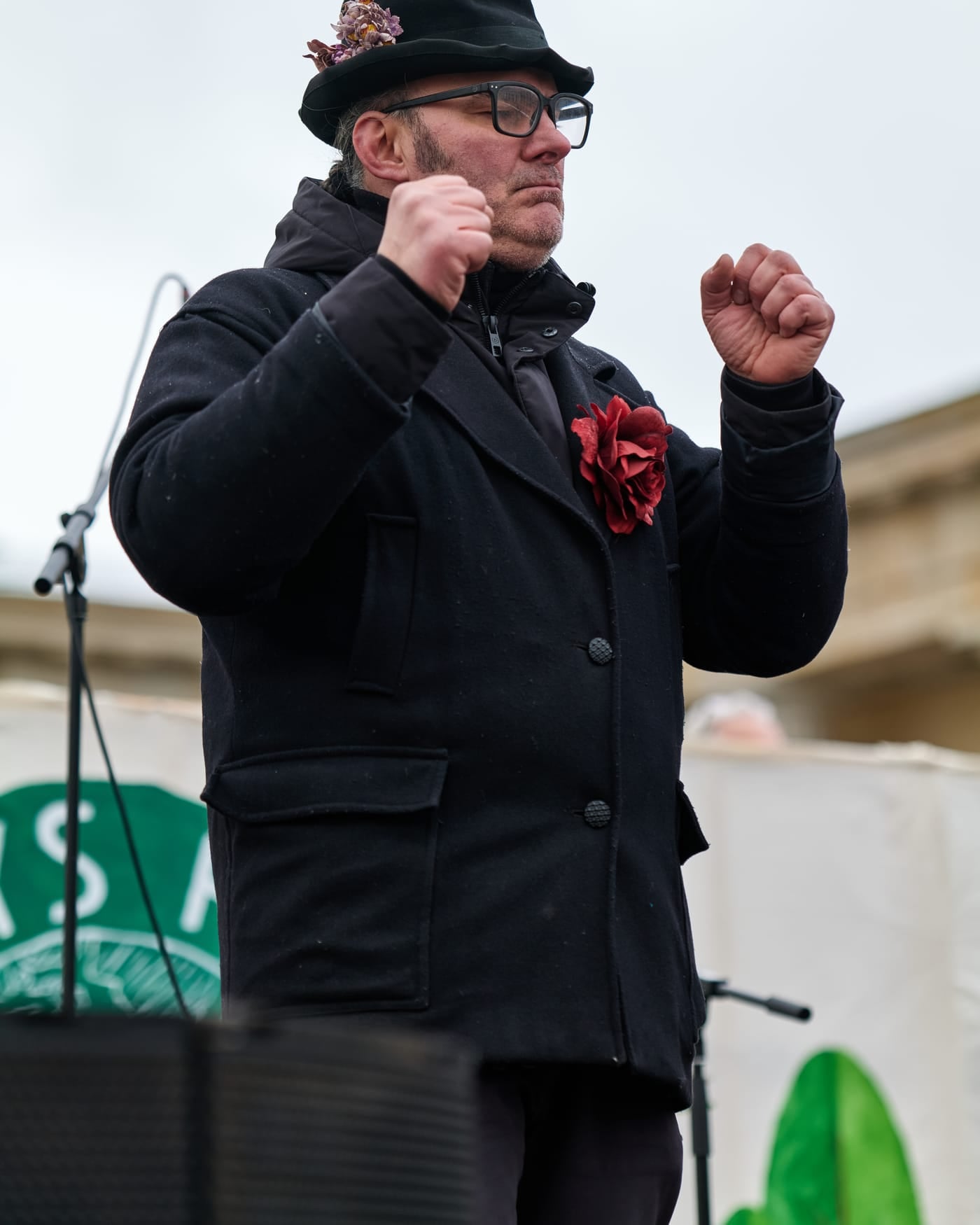 Sign language interpreter in a dark coat and hat signing with both hands at an outdoor event