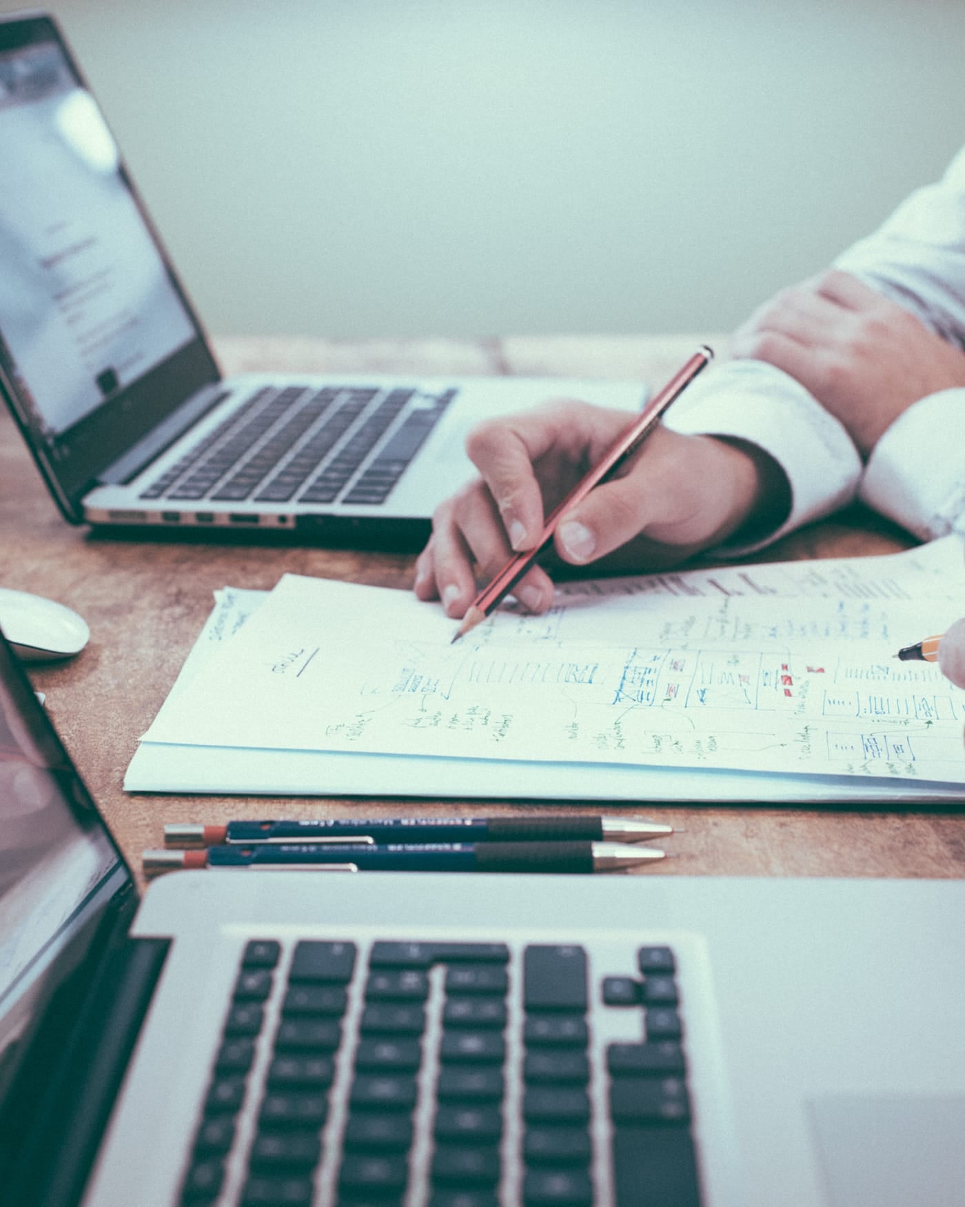 Hands writing notes on paper between two open laptops at a wooden meeting table