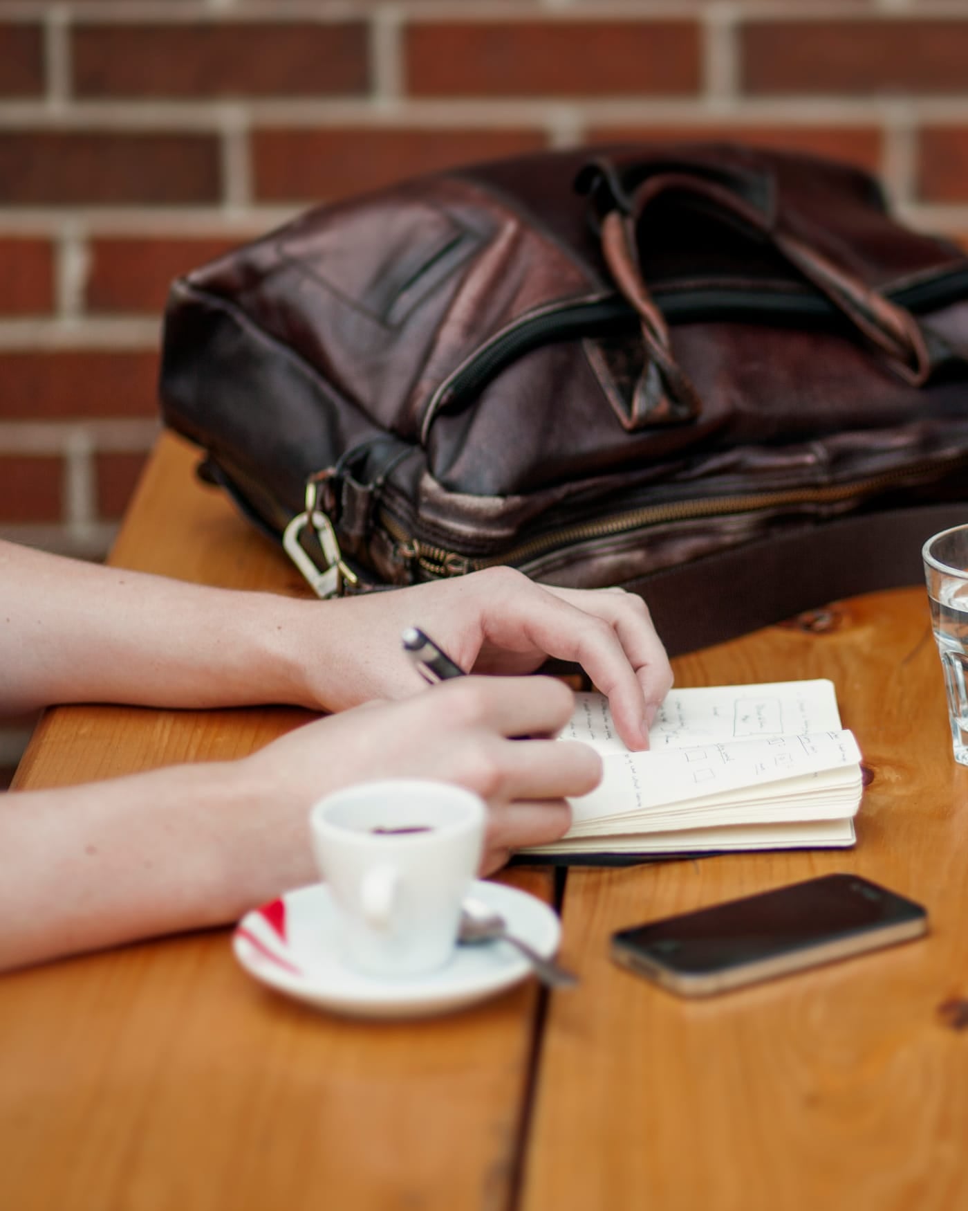 Hand taking notes in a small notebook at a wooden table with an espresso cup, phone and leather bag