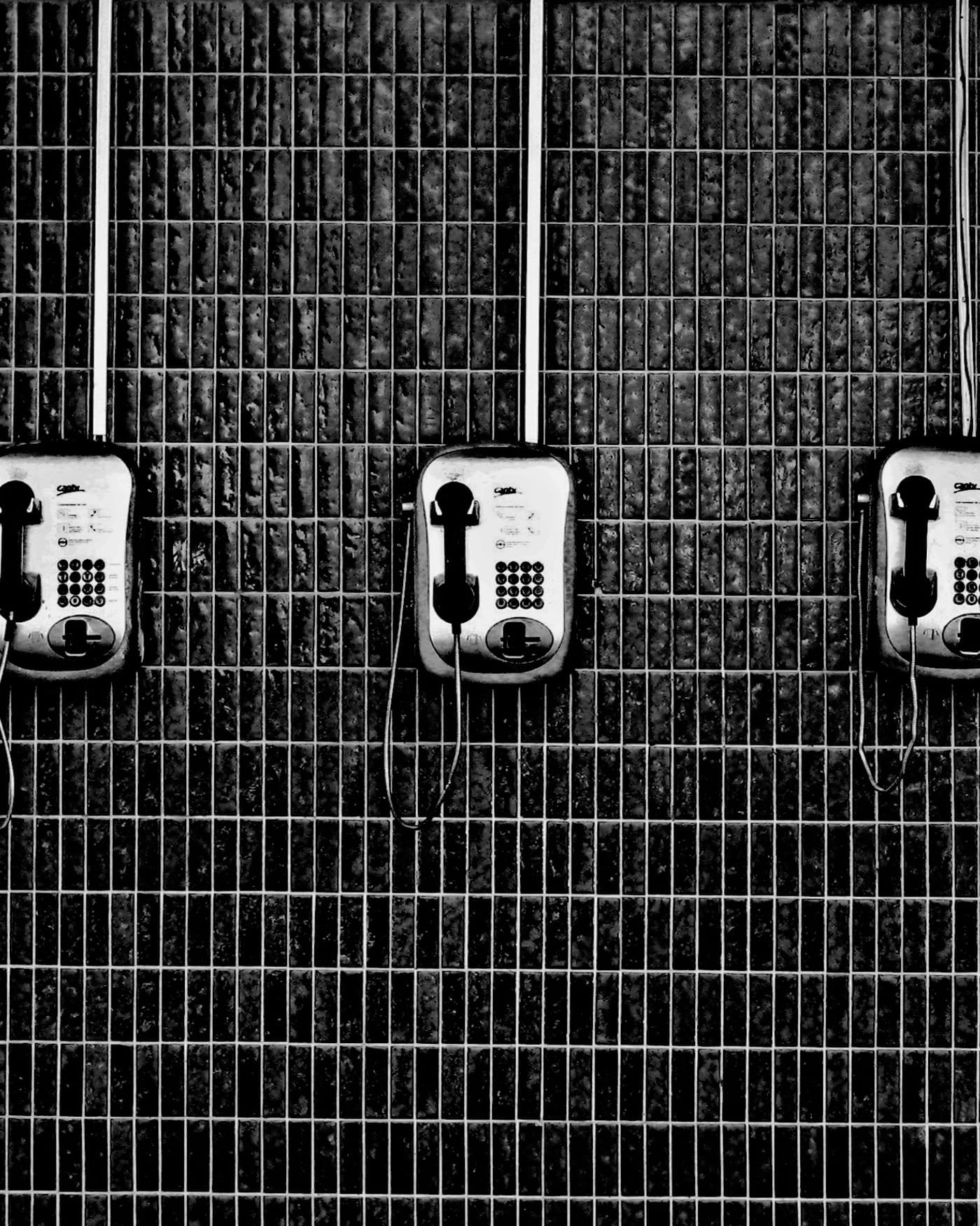 Row of three wall-mounted payphones on a dark tiled wall, black and white