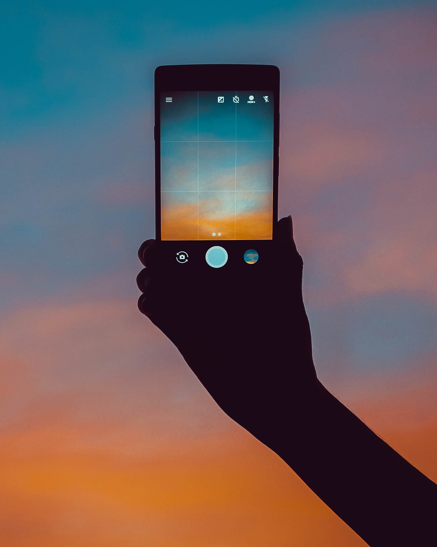 Silhouetted hand holding up a smartphone against an orange and blue sunset sky