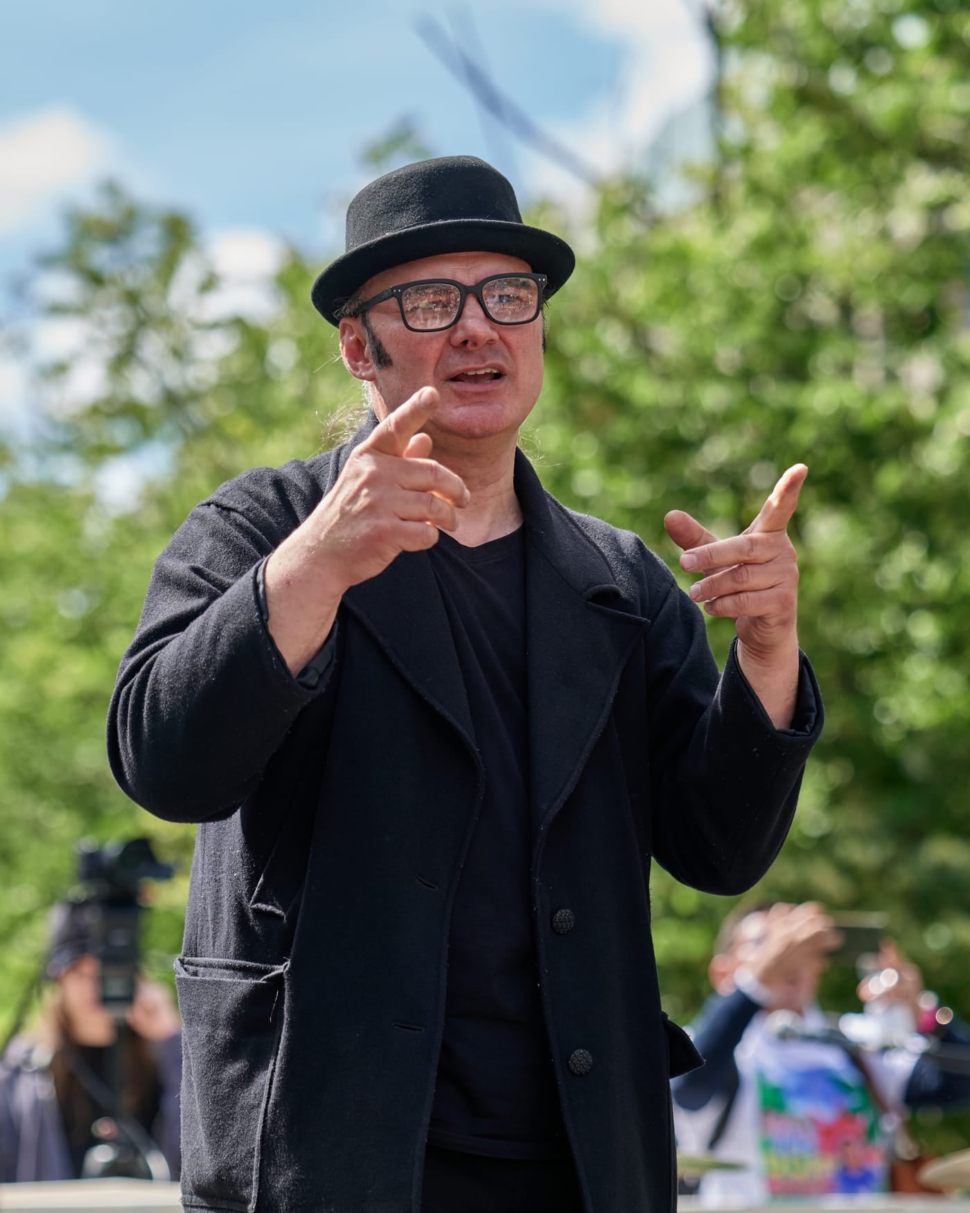 Sign language interpreter in a black coat and hat signing on an outdoor stage in front of green trees
