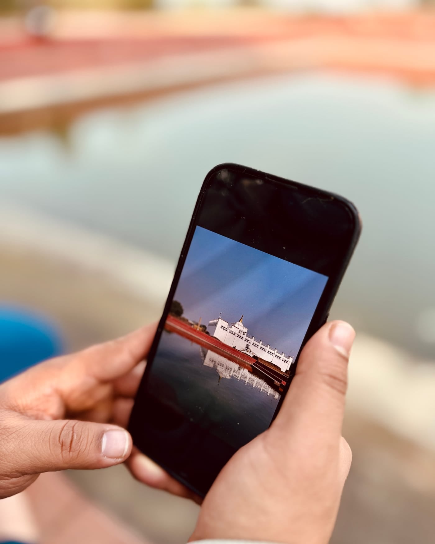 Two hands holding a black smartphone that shows a photo of a white temple reflected in a pond