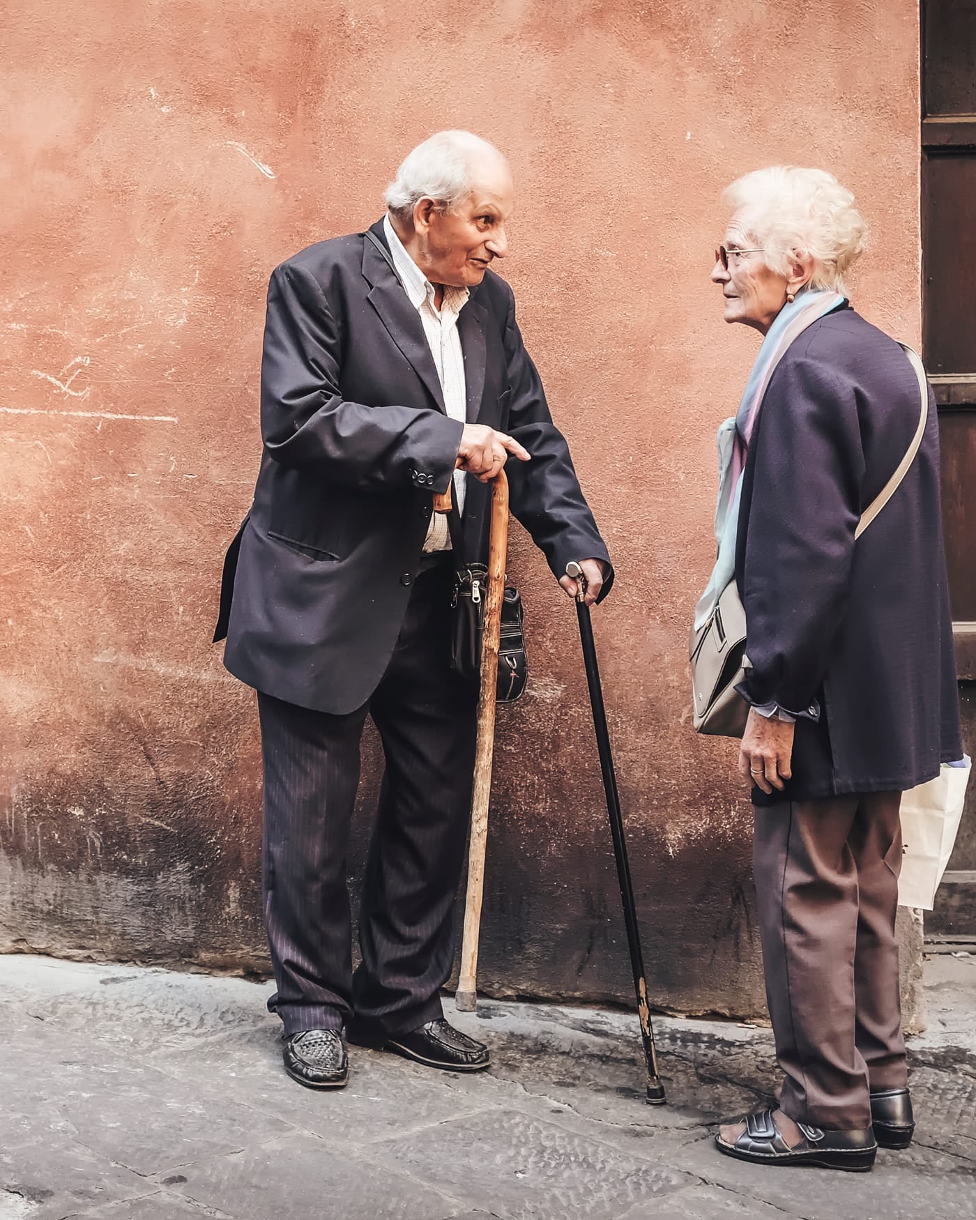 Two elderly men in dark suits talking on a street beside a terracotta wall