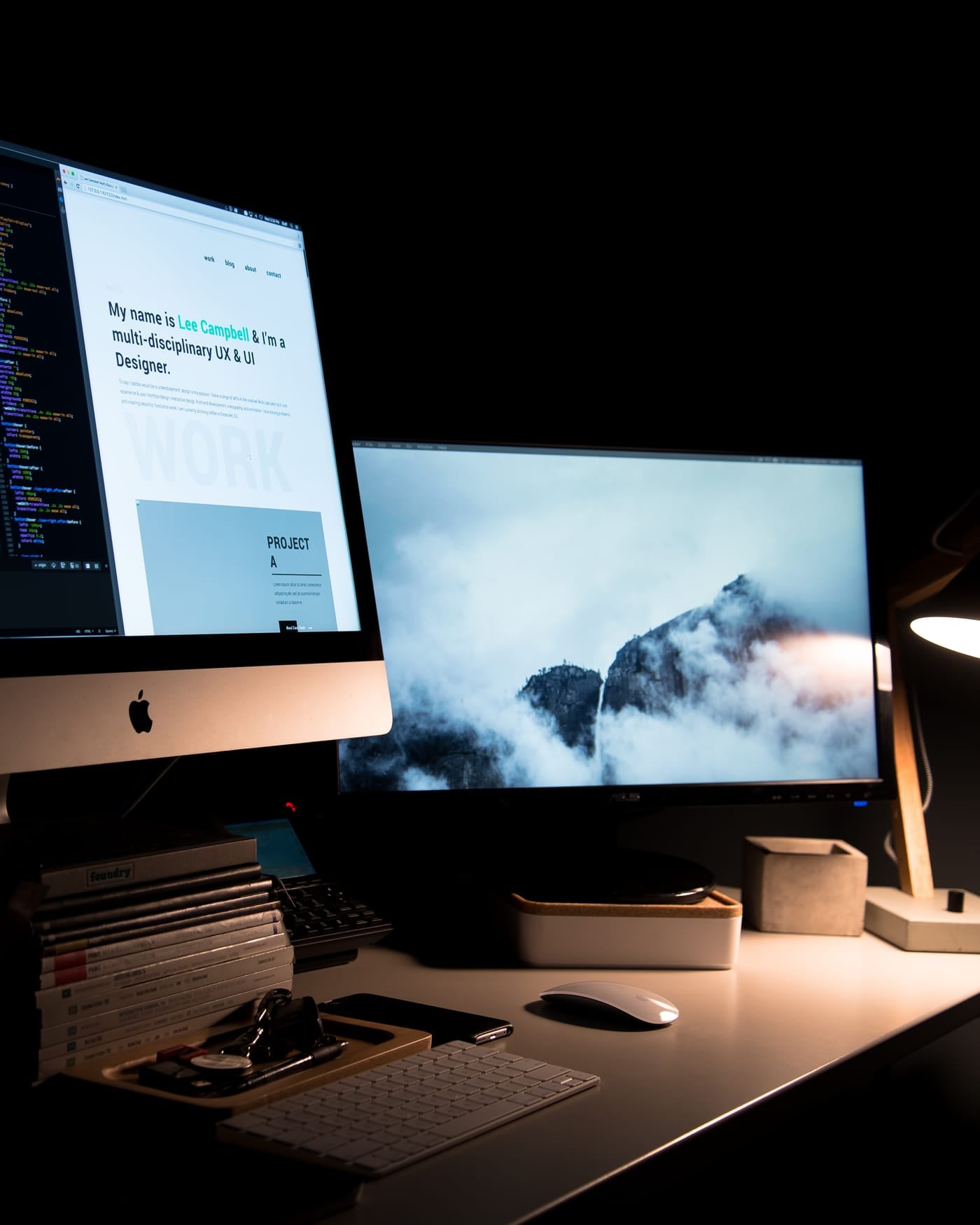 Dark desk with an iMac showing code and a second monitor with a mountain wallpaper under a lamp